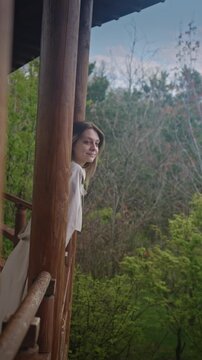 Woman leaning from wooden balcony of cabin overlooking forest, travel writer pausing in morning light, gentle breeze and green