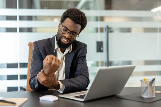Business man sitting at a desk and holding his painful wrist, suffering from an injury related to repetitive strain and long hours working on a laptop in a modern office environment