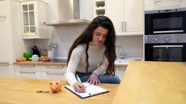 Young woman sitting on a kitchen counter, diligently writing on a clipboard to manage her budget, with a piggy bank and coins placed next to her, symbolizing financial planning and savings