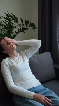 Dizzy young female headache or migraine pain suffering from vertigo or sick, holding head with hands, health problem of brain or inner ear not balance, sitting alone on couch at home