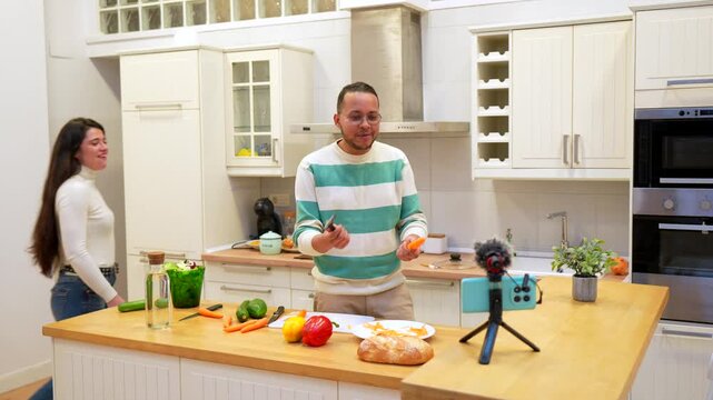 Young man vlogging about cooking healthy food while his girlfriend surprisingly joins him, hugging, dancing, and laughing together in front of the smartphone camera on a tripod for a live stream
