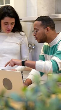 Happy diverse couple reviewing documents and using a laptop in their kitchen, managing household finances, paying bills online, and planning their budget with a positive and collaborative attitude
