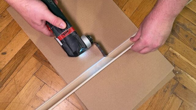 Hands use a cordless drill to drive a screw into a cardboard sheet on a wooden floor, practical home repair scene with focused do it yourself mood