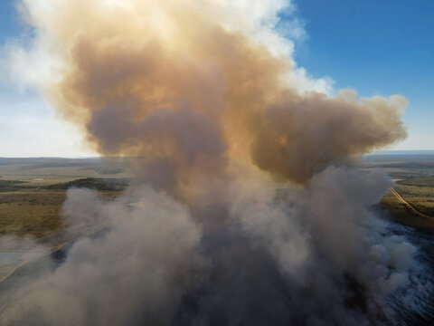 Aerial view of thick plumes of smoke rising from a wildfire over the vast savanna landscape under a clear blue sky in Waterberg District Municipality, Limpopo, South Africa.