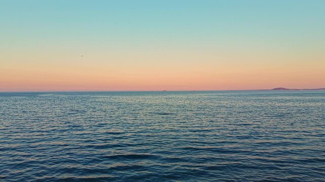 A sunset occurs over the ocean, casting colors across the sky. The water reflects hues from the sky. Land appears in the distance, creating a peaceful scene by the shore.