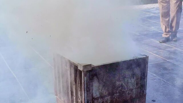 A rusted metal burn bin is engulfed in thick, billowing beige smoke that obscures the flames inside, as people stand nearby on the tiled ground monitoring the fire�s suppression.