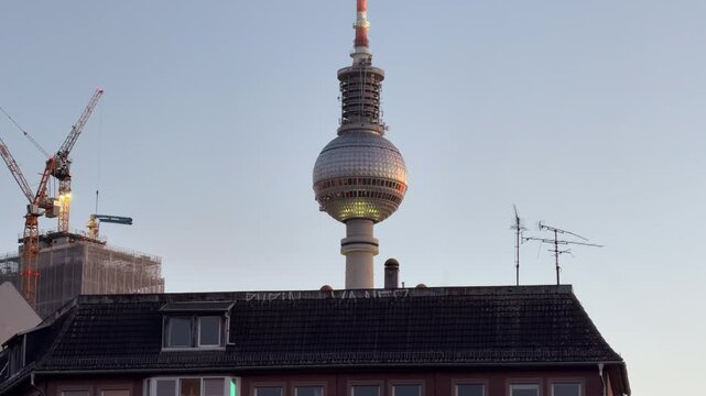 Handheld shot of Berlin TV Tower, Fernsehturm, rising above city buildings and cranes in Alexanderplatz at sunset, symbolizing urban development