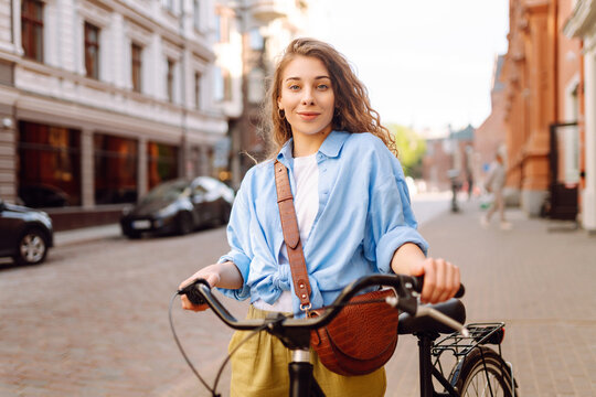 A woman holds her bicycle in a city square while smiling. People walk by in the background, and buildings stand tall. The weather is clear and sunny. Lifestyle.