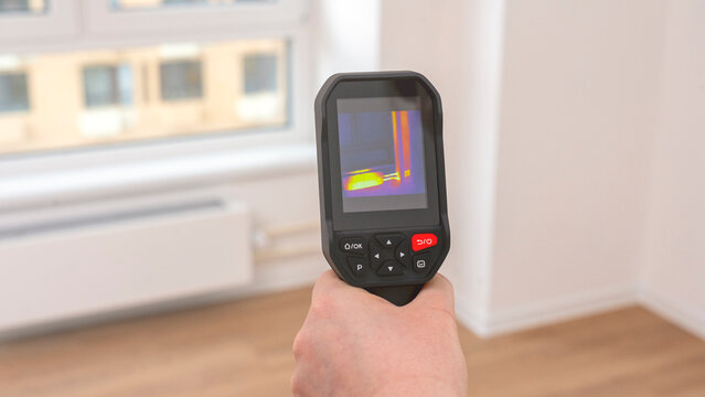 Hand holds a thermal imaging camera. Its screen shows color-coded temperature patterns. The background features a window and radiator. This image applies to home energy audits