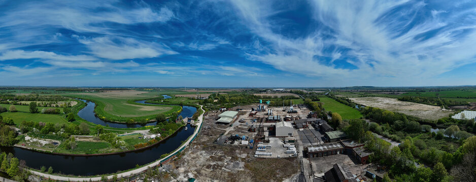 Aerial view of an industrial complex situated beside a winding river and lush green meadows under a blue sky with wispy clouds in Knottingley, England, United Kingdom.
