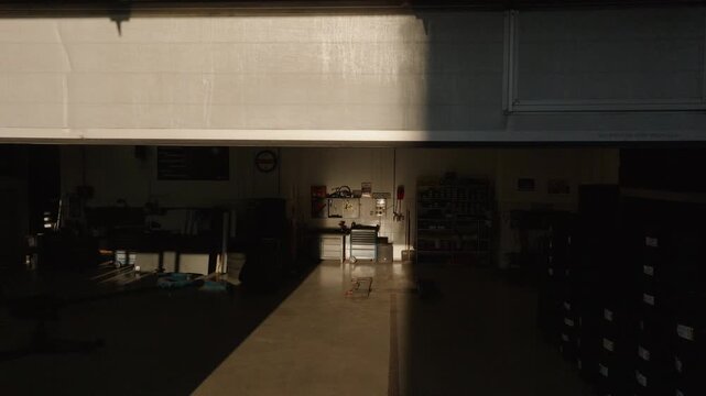 Workshop beginning its working hours, roller door lifting, and the sunlight illuminating a dark professional car repair shop, drone shot.