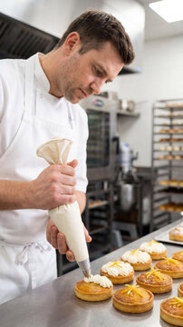 Middle-aged Caucasian male chef in a white uniform piping cream onto fruit tarts in a professional bakery. Focused craftsman working in a commercial kitchen with ovens behind