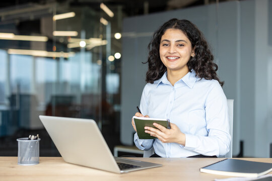 Smiling indian businesswoman sitting at desk in a modern corporate office, holding a notebook and pen while looking at the camera, with a laptop and organized workspace