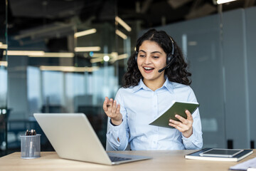 Fototapeta premium Young woman wearing a headset and holding a notebook, having a virtual meeting on her laptop while providing customer support from a modern office environment