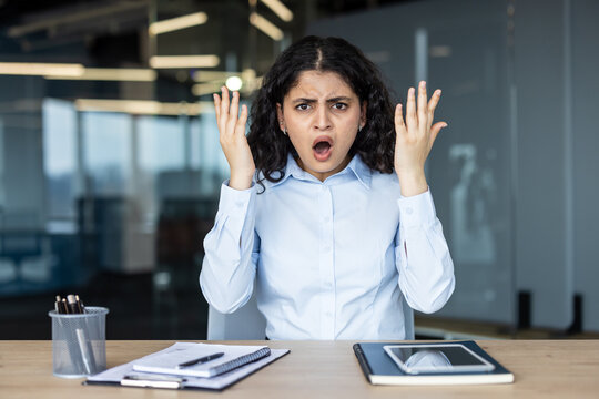 Young businesswoman with dark curly hair shows shock and frustration, hands raised at desk in a modern office, overwhelmed by stress and exasperation during a work crisis