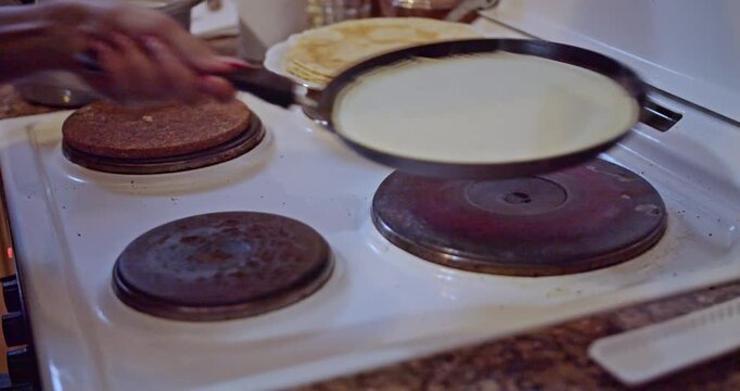 A close-up view of hands skillfully flipping a thin crepe in a hot pan on an electric stove. A stack of golden, freshly cooked crepes sits nearby, ready for a delightful homemade meal.