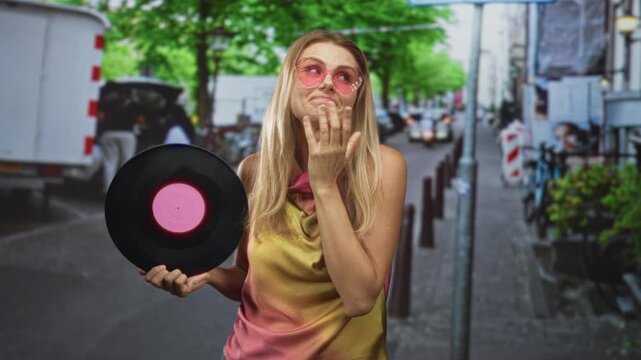 Woman holding a vinyl record with left hand while right hand touches cheek on a street, wearing pink sunglasses and a sleeveless top; nostalgia memory retro.