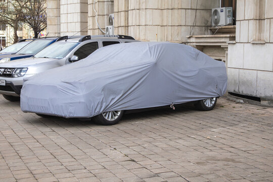 A sedan car parked on a paved sidewalk completely covered with a protective gray fabric tarpaulin - Bucuresti, Romania - March 15 2026