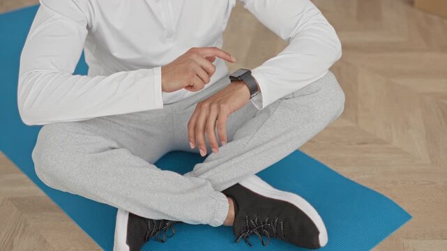 Man touching smartwatch on wrist while seated crosslegged on blue yoga mat in studio wearing grey sweatpants and black sneakers; calm focus.
