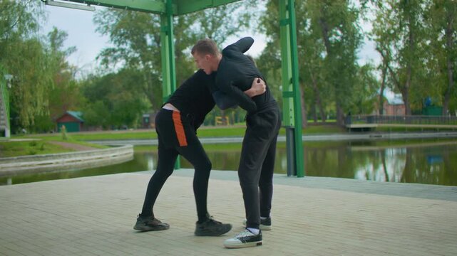 two white men grappling by pond under green pavilion, coach guiding student through takedown sequence with close clinches, gripping and footwork, autumn trees reflecting in water, focused expressions