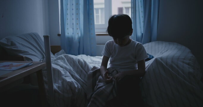 Boy sitting on bed in dark bedroom with head down, conveying sadness, melancholy, and signs of childhood depression