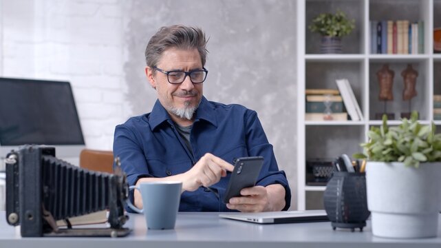 Bearded man working online with laptop computer and smart phone at home sitting at desk. Businessman in home office. Portrait of mature age, middle age, mid adult man in 50s.