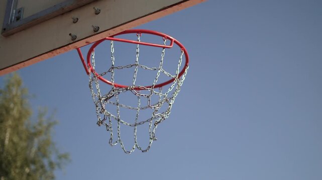 Basketball ball hitting rim of hoop, bouncing and falling into metal net, gimbal shot from behind basketball backboard. Ball landing exactly on target. Winning throw without players outdoors.