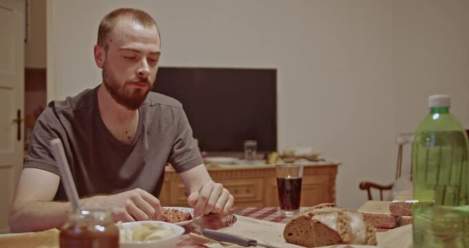 A young bearded man sits thoughtfully at his dining table, enjoying a simple meal alone. The quiet domestic scene captures a moment of calm reflection during an everyday activity at home.