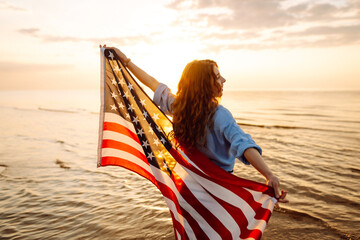 A cheerful woman stands on the seashore with an American flag at sunset. A young woman walks along the beach holding a flag. Holiday concept, patriotism. Independence Day. © maxbelchenko