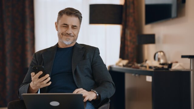 Content middle-aged man multitasking with phone and laptop. Businessman working at home with computer, smiling. Happy mid adult male with content smile. Entrepreneur managing business online.