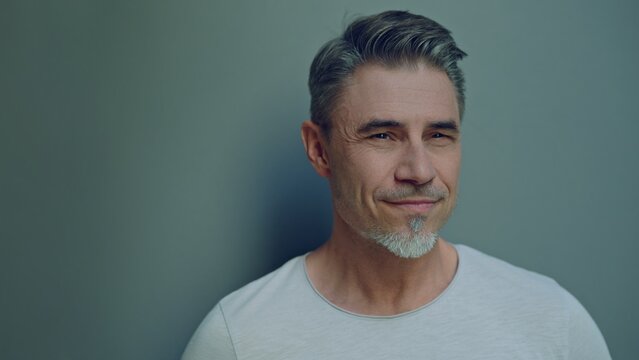 Close-up portrait of a smiling middle-aged man with a grey beard, against a soft gray background. Happy mid adult male in 50s, bearded, grey hair, confident friendly smile, looking away.