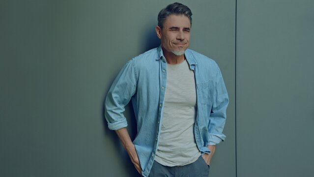 Portrait of a smiling man in 50s with a relaxed, casual look. Happy male standing hands in pocket. Blank copy space for text on grey wall background.