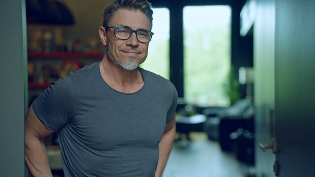 Portrait of smiling man in 50s wearing glasses and casual grey t-shirt, exuding confidence and poise in a modern indoor setting. Happy mid adult, middle aged male with satisfied confident smile.