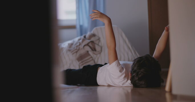 Boy lying on bedroom floor with arm raised in air, appearing bored and restless, symbolizing childhood idleness, lack of engagement, and waiting in silence indoors