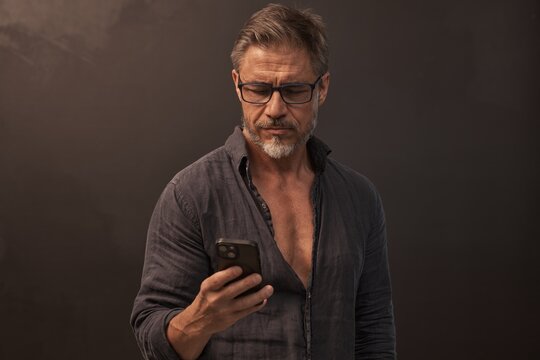 Portrait of mid adult man standing against dark background in gray casual clothes. Studio shot of middle aged male in glasses using mobile phone.