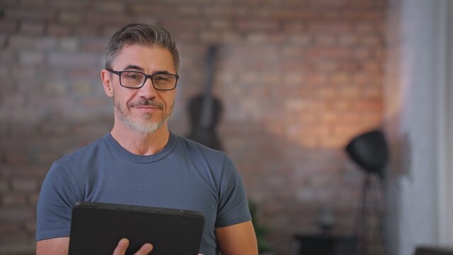 Middle aged man using digital tablet at home in loft living room. Happy, sporty, active senior male with gray hair and glasses reading social media, working and smiling.