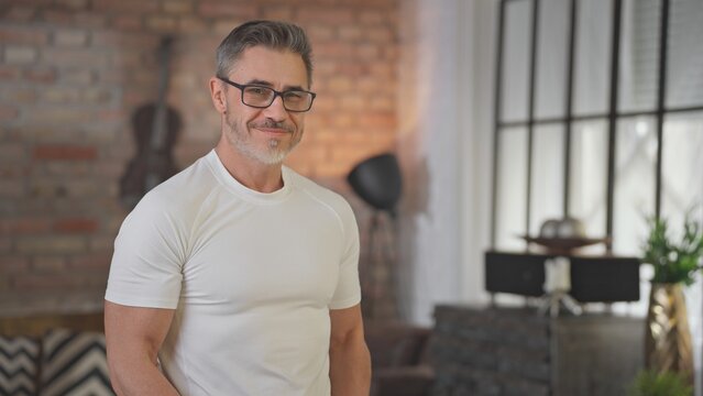 Portrait of happy confident casual middle aged man at home in loft room. Fit and healthy active senior male with grey hair looking at camera, smiling. Books in background, smart older guy.