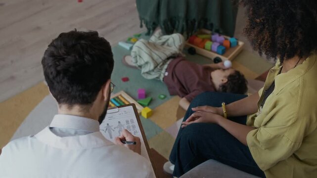 High angle view of young Black mother communicating with pediatrician filling out medical card of toddler patient playing with toys on floor during home appointment