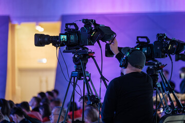 Cameraman operates professional video camera on tripod during live conference, colorful stage...