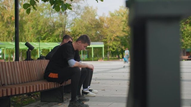 two young men sitting on bench, urban park plaza with autumn leaves, one hunched forward with head in hands while companion looks distant, quiet mood of solitude and contemplation, slow camera pan