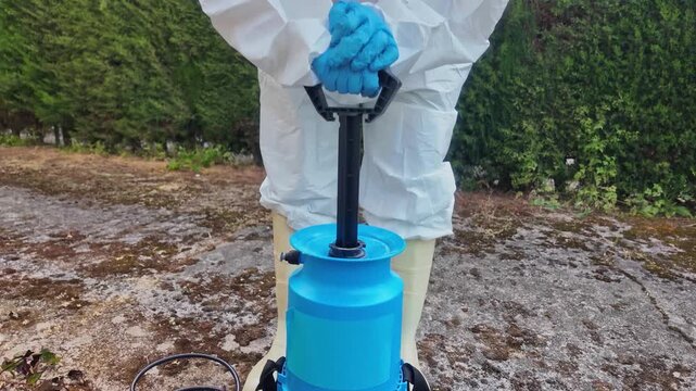 Person in protective suit operates manual pump to pressurize blue spray tank for effective pesticide application in garden.