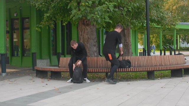 two young men sitting on bench outside green building, student and courier roles implied, backpack on ground, casual black clothing, one scrolling phone while other watches and gestures, autumn leaves
