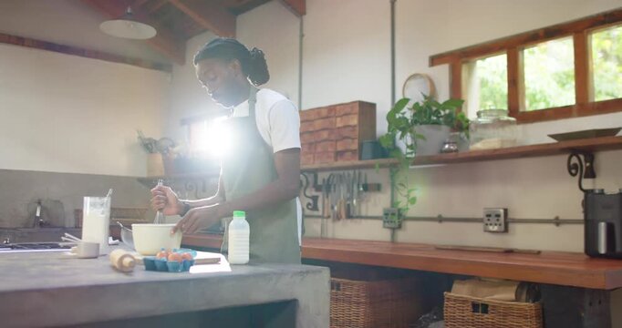 African American man in green apron whisking batter at island, sun flaring causing recipe demo