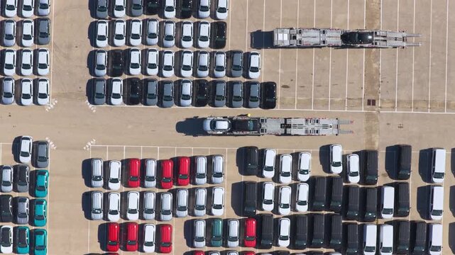 Numerous new vehicles neatly arranged in a large parking facility as car transporter trucks maneuver, showcasing the automotive industry's manufacturing, sales, and logistics process