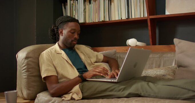 African American man starting typing on laptop on leather couch, pausing to think, bookshelf