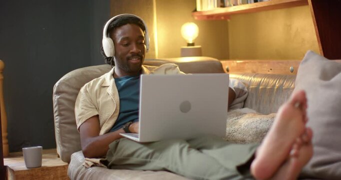 African American man lounging and typing on laptop on sofa for work, camera panning revealing lamp