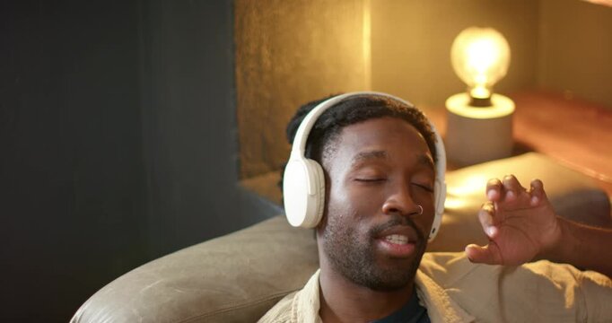 Man reclining at home, adjusting white headphones and mouthing lyrics to music playing