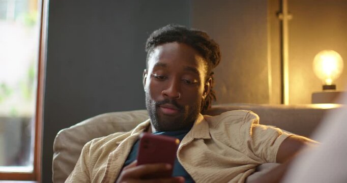 African American man sitting on couch at home, glancing at window, returning to red phone, smiling