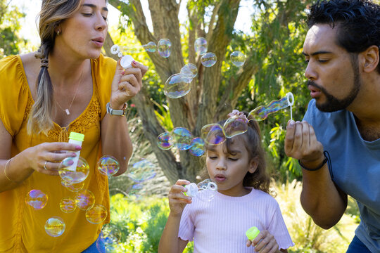 Diverse family blowing bubbles with neon-capped bottles and wands in sunny park wearing mustard-top