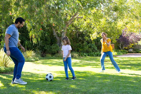 Parents with youth girl playing with soccer ball on sunlit lawn under tree, woman approaching